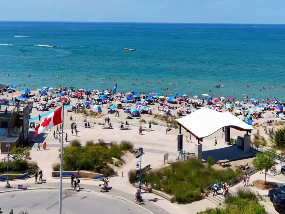 Grand Bend Main Beach from above on a summer day: umbrellas along the sand, the pavilion by the splash pad and the blue water of Lake Huron