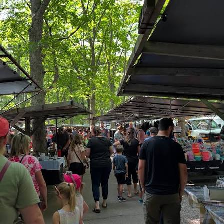 Shoppers walking between vendor tents under the trees at the Pinery Market