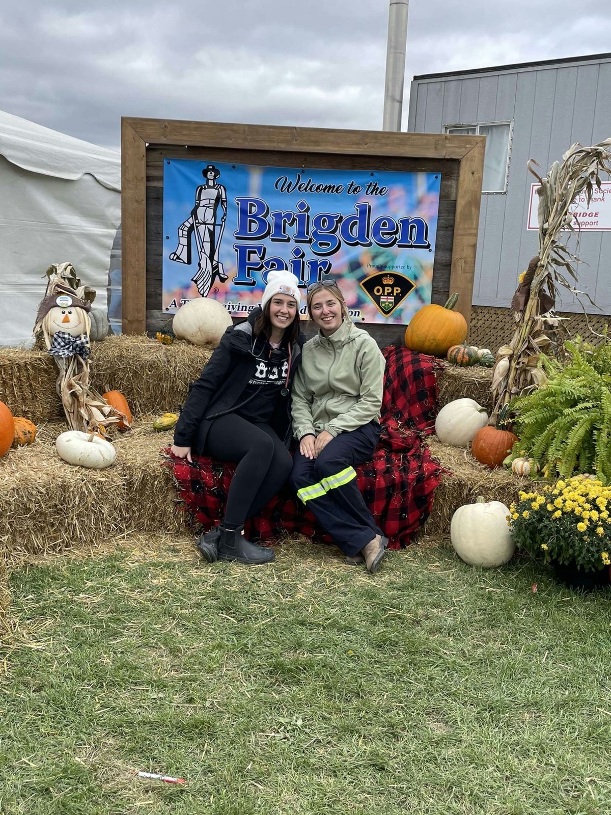 Two first responders sit on hay bales in front of the Welcome to the Brigden Fair sign, surrounded by pumpkins and mums.