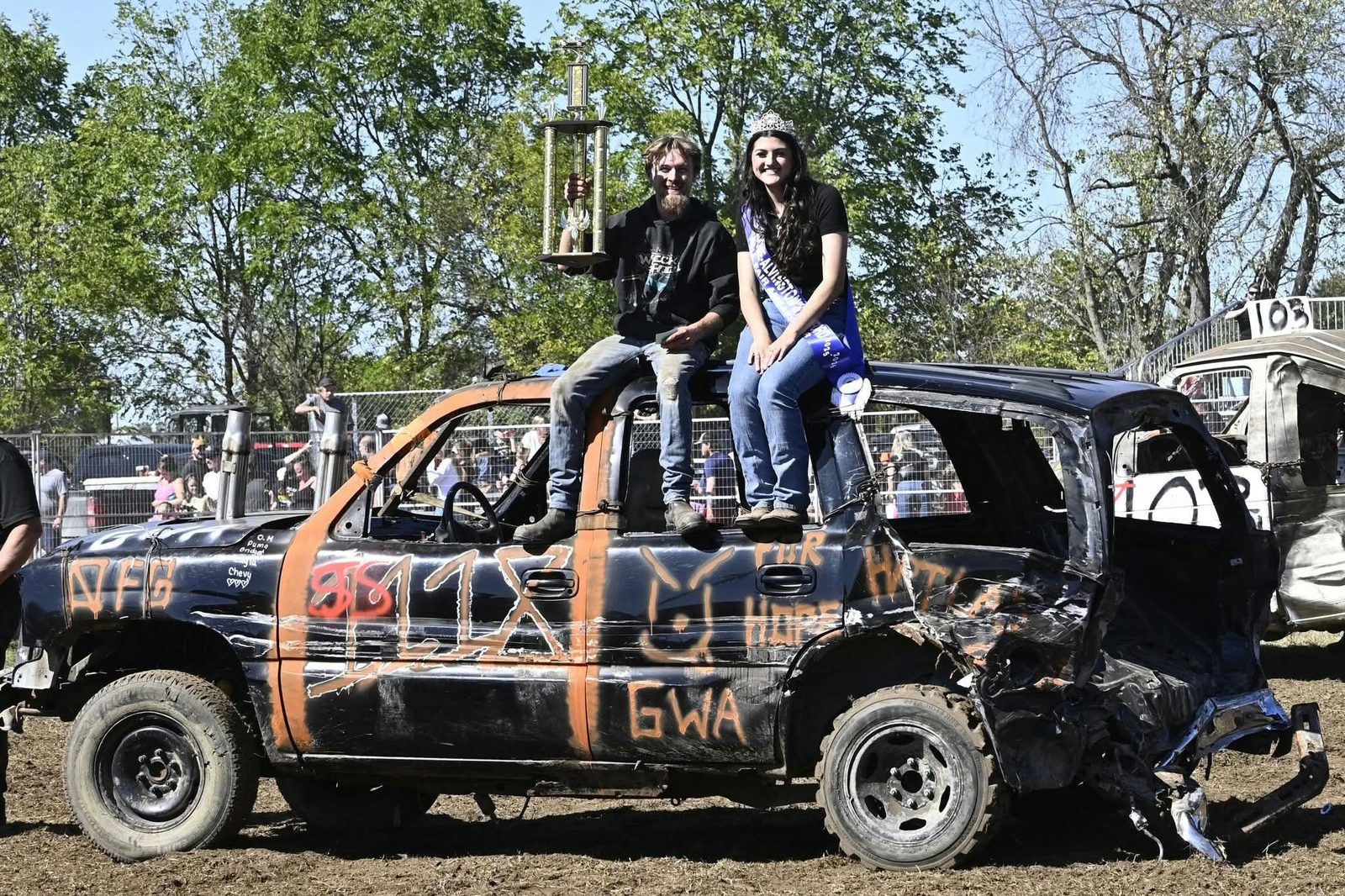 A demolition derby winner and the fair ambassador sit on the roof of a battered car holding a trophy.
