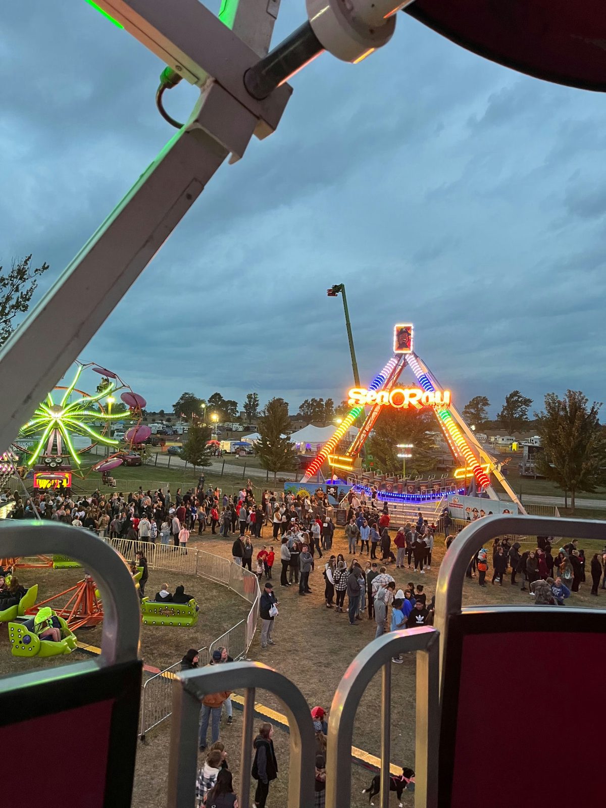 The view from inside a ferris wheel car at dusk, looking down over the lit midway and the crowd below.
