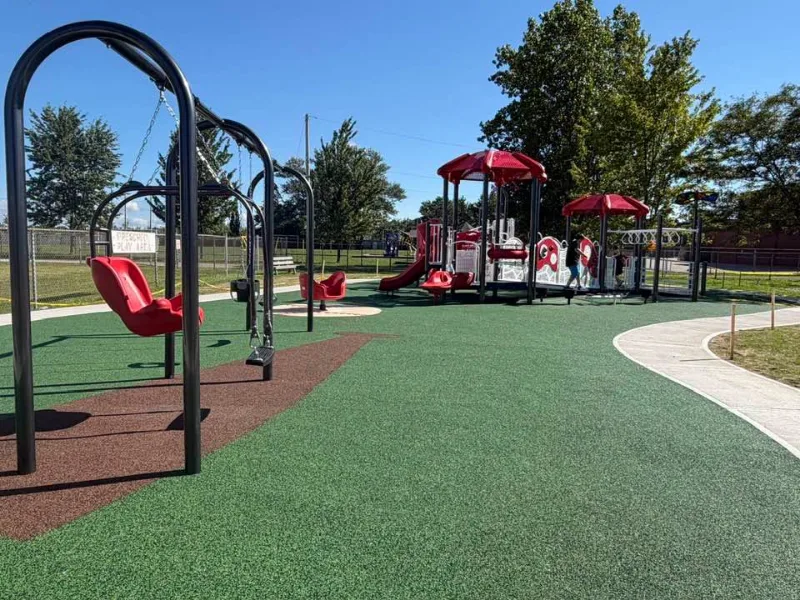 The playground at Centennial Park in Watford, with red swings and a red-roofed climber on green rubber surfacing
