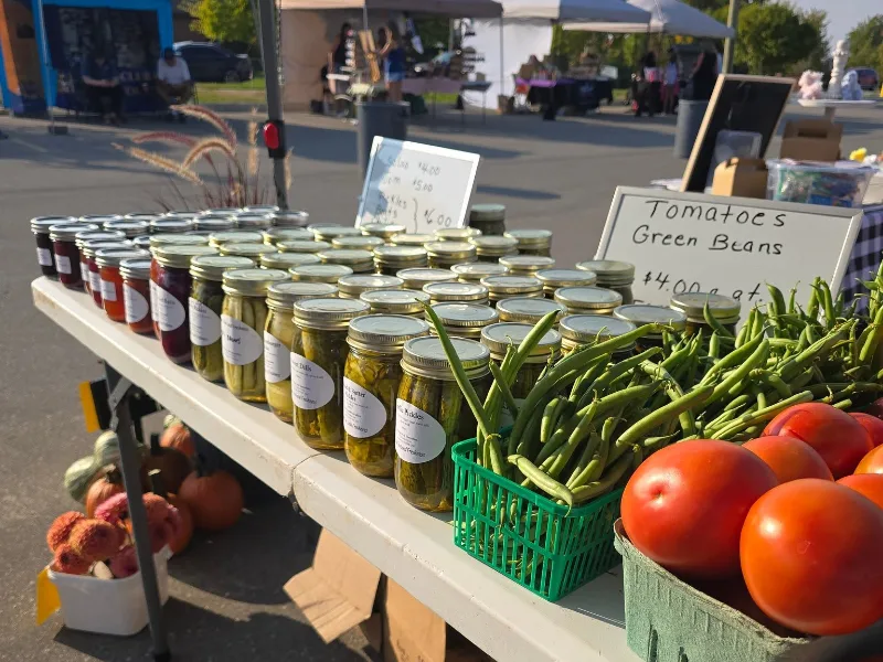 A market table of pickles, preserves, green beans and tomatoes, with vendor tents behind