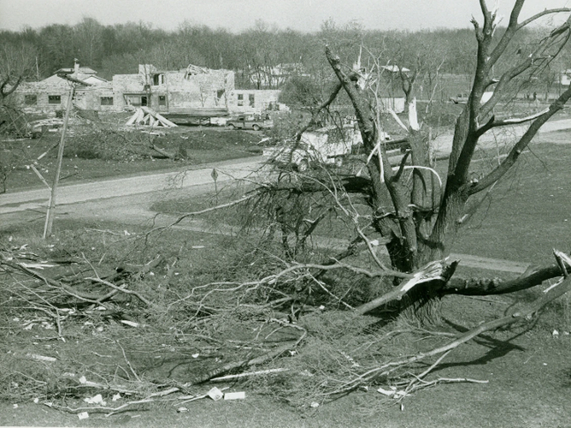 A black-and-white photograph from May 1983 of a building torn open to its walls and trees snapped along the tornado's path