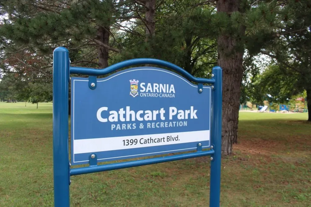 The blue City of Sarnia Parks and Recreation sign for Cathcart Park at 1399 Cathcart Blvd, with the play area visible through the trees