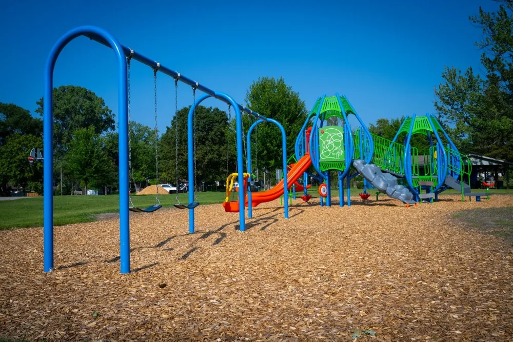 The blue swing set and bright green play structure at Harry Turnbull Park in Sarnia, on a wood chip surface under a clear sky