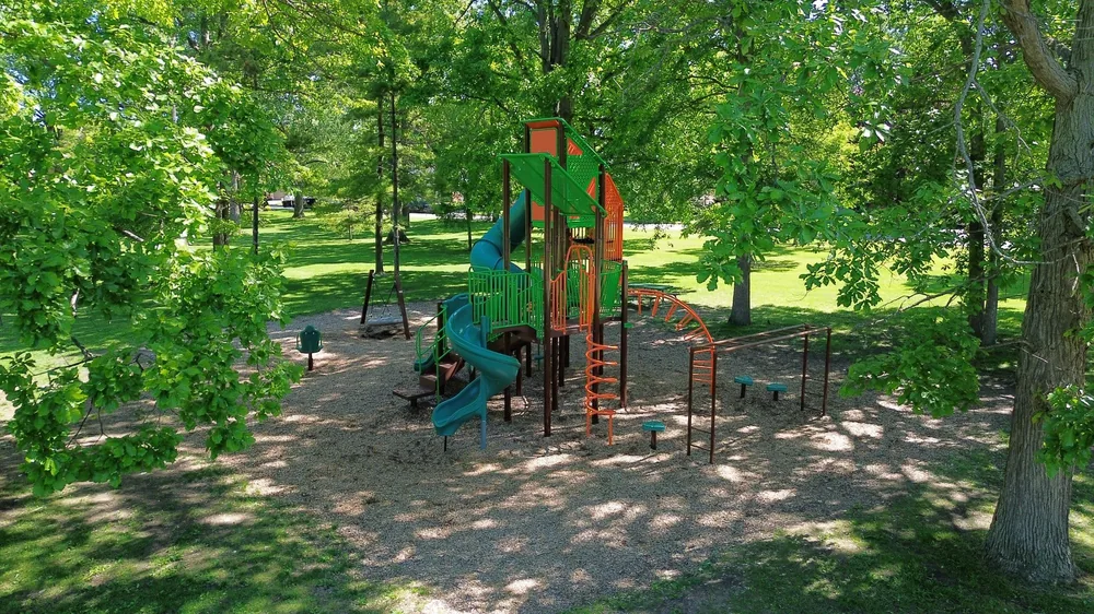 The tall green and orange tower structure at Marcin Park in Sarnia, with a spiral slide and climbing bars, shaded by tall trees