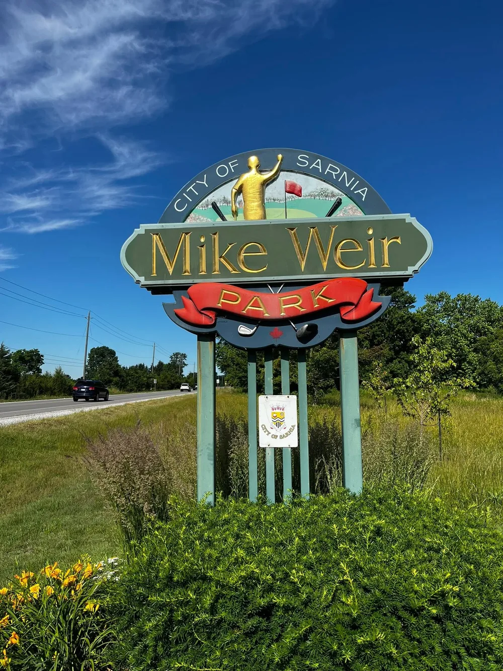 The City of Sarnia entrance sign for Mike Weir Park on Lakeshore Road, with a golfer emblem and wildflowers at its base