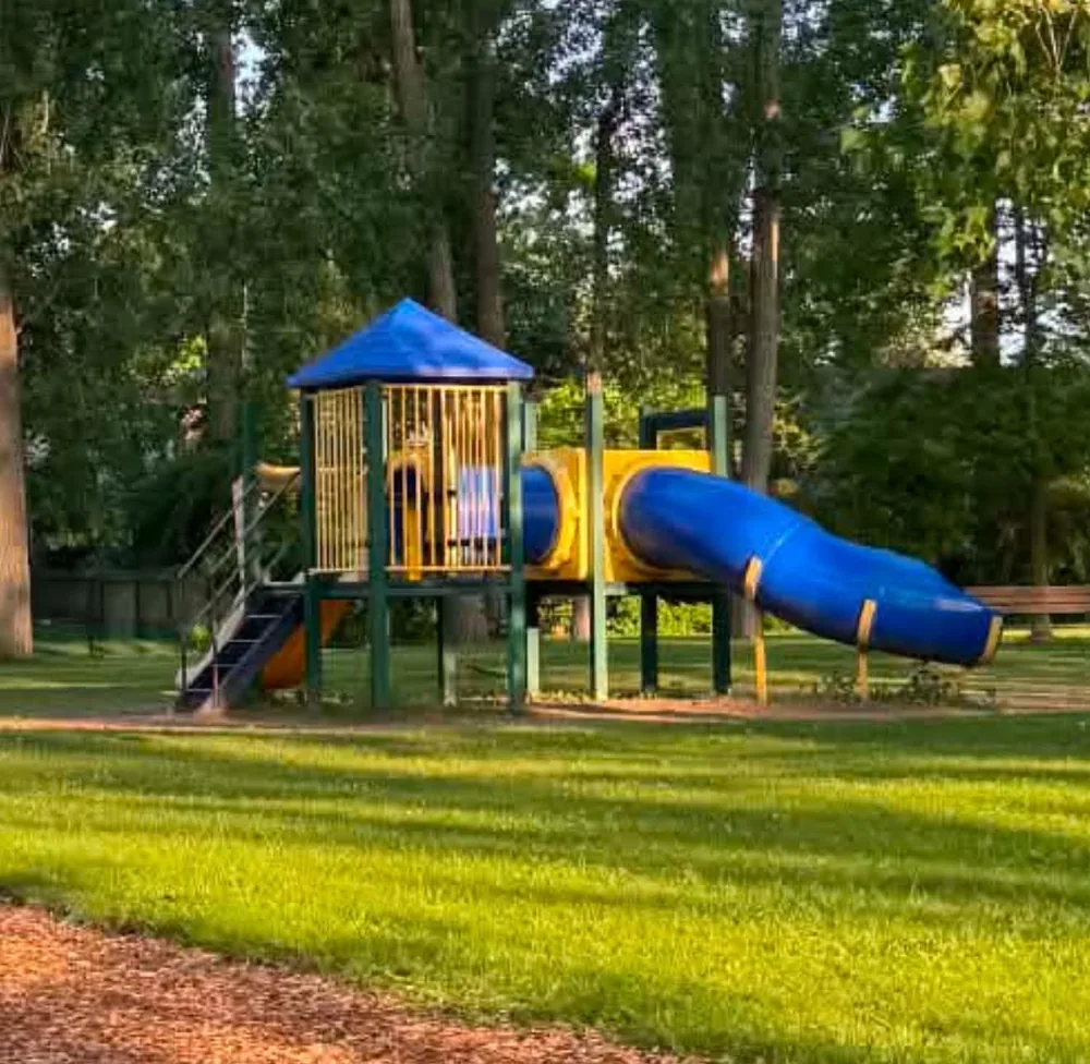 The older green and yellow tower structure at Twin Lakes Park in Sarnia, with a blue enclosed tube slide, surrounded by mature trees and lawn