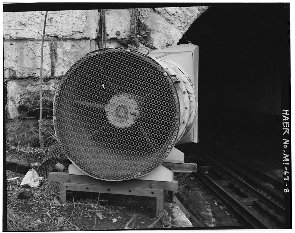 A large mesh-guarded exhaust fan mounted on a steel stand beside the rails at the mouth of the stone tunnel portal, the dark bore opening alongside it